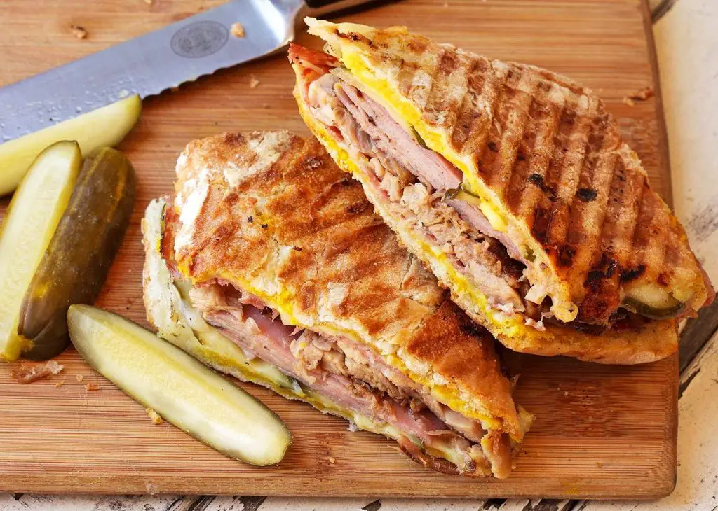 A Cuban sandwich cut in half with a pickle on a cutting board.