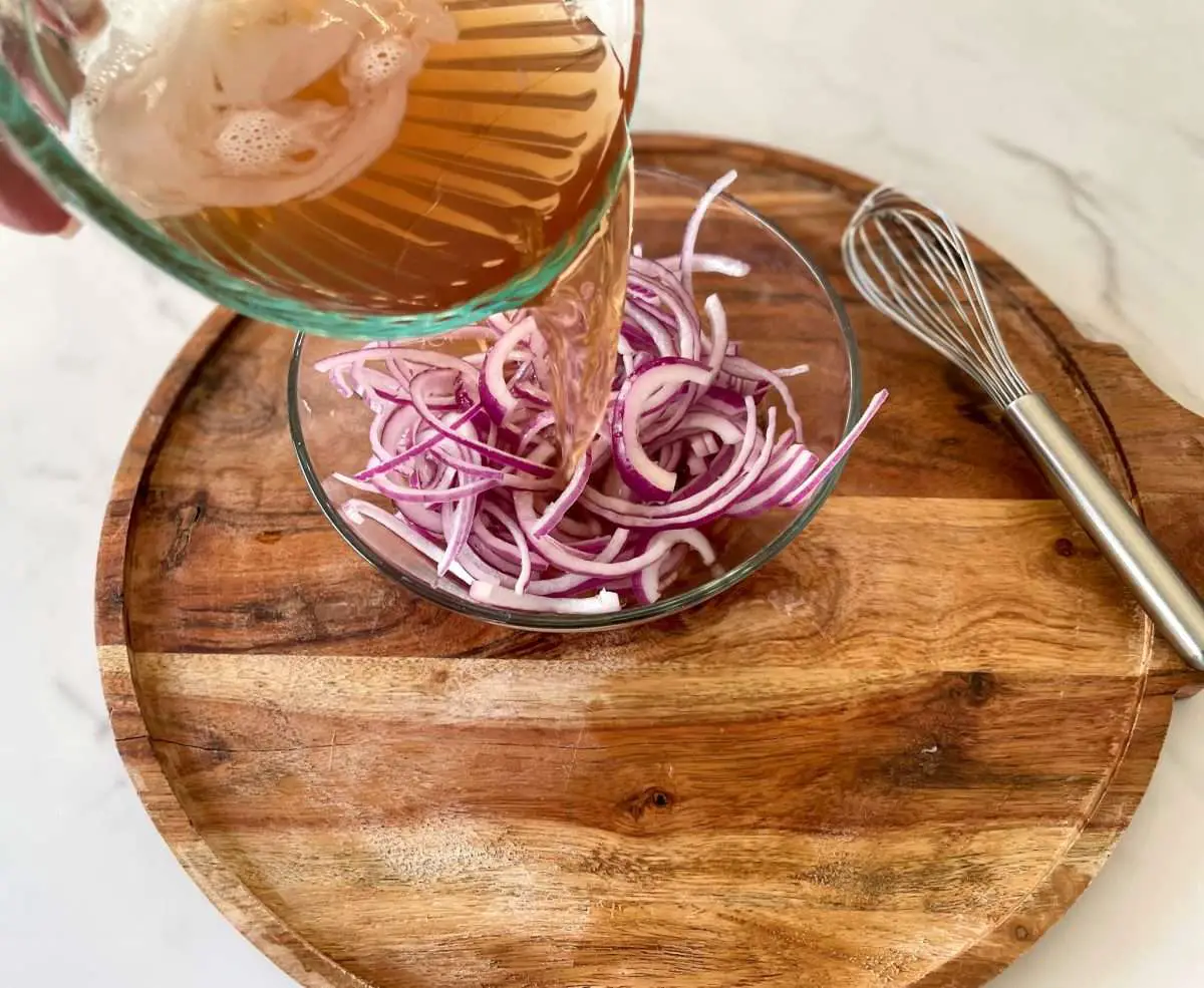 Pickling liquid being poured over thinly sliced red onions on a board.