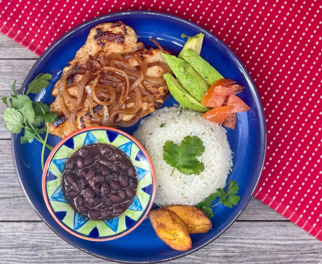 Pollo a la plancha, white rice, black beans, maduros and avocado on a blue plate.