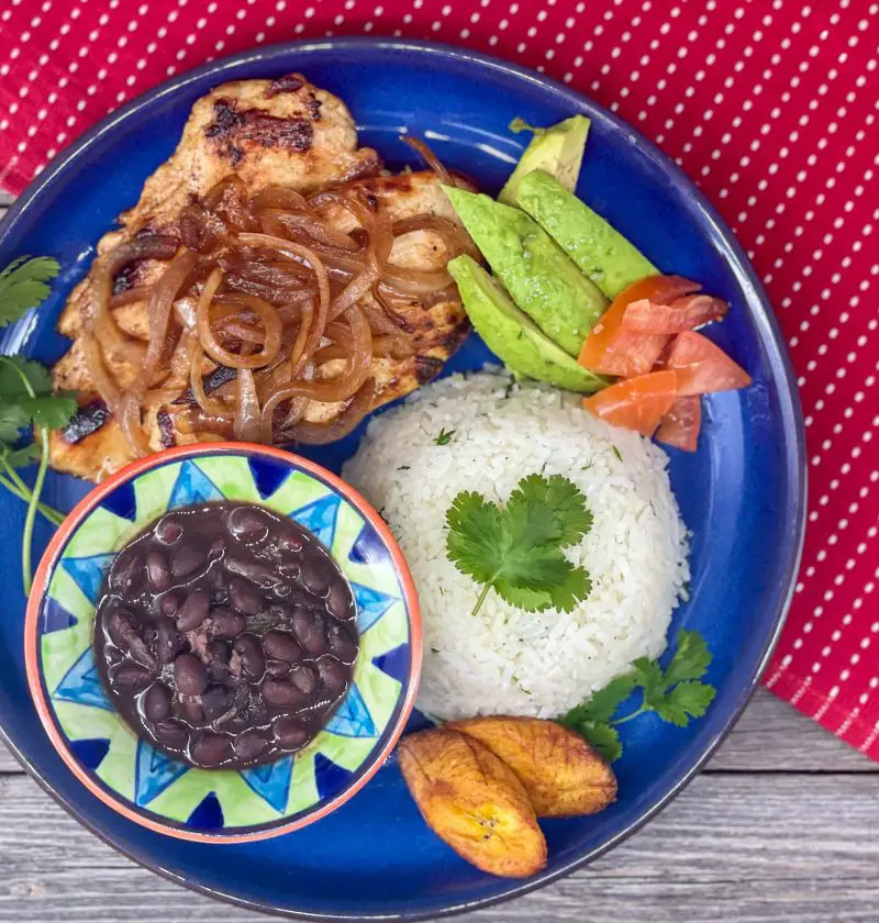 Pollo a la plancha, white rice, black beans, maduros and avocado on a blue plate.